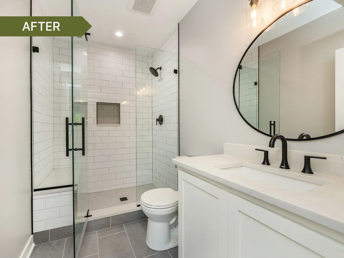 Modern bathroom with white subway tile shower, glass enclosure, black fixtures, round mirror, and white vanity with quartz countertop.