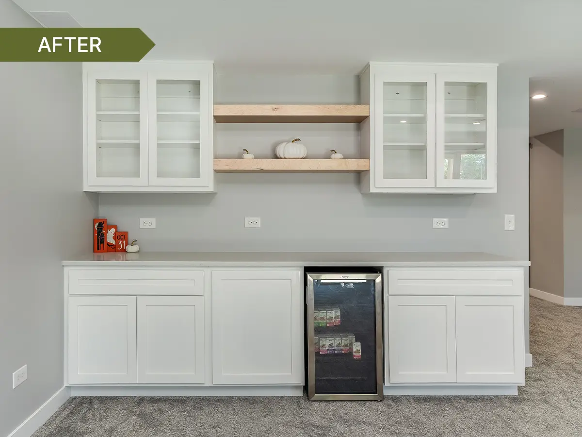 Finished basement wet bar with white cabinets, glass-front uppers, floating wood shelves, gray countertop, and a built-in beverage fridge.