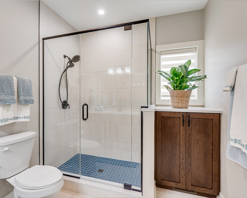 Modern bathroom with glass walk-in shower, blue tile shower floor, beige wall tiles, wood vanity cabinet, window with zebra blinds, and towel rack beside the toilet.
