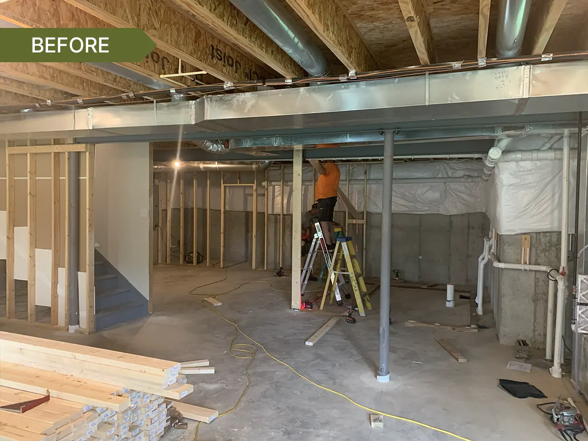 Unfinished basement with exposed framing, ductwork, support beams, and a worker on a ladder during construction.