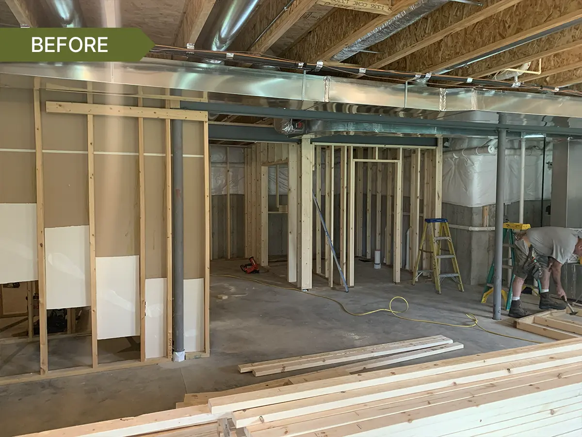 Unfinished basement under construction with exposed framing, ductwork, support beams, concrete floor, and lumber stacked in the foreground.