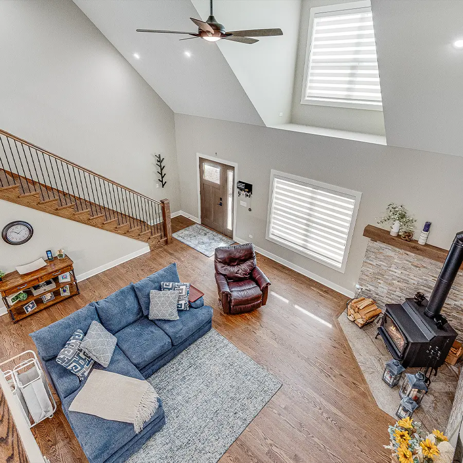 Open living room with vaulted ceiling, staircase railing, blue sectional sofa, leather recliner, wood floors, and a stone fireplace with a wood-burning stove.