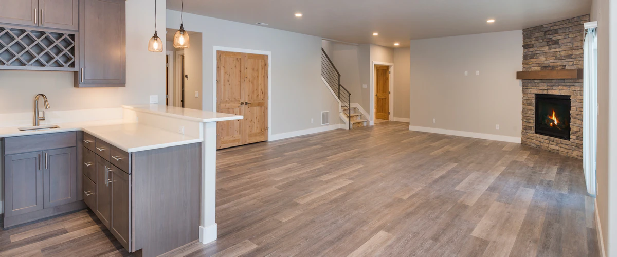 Large finished basement area with wood-look flooring, a stone-wrapped gas fireplace, and a partial view of a gray kitchen.