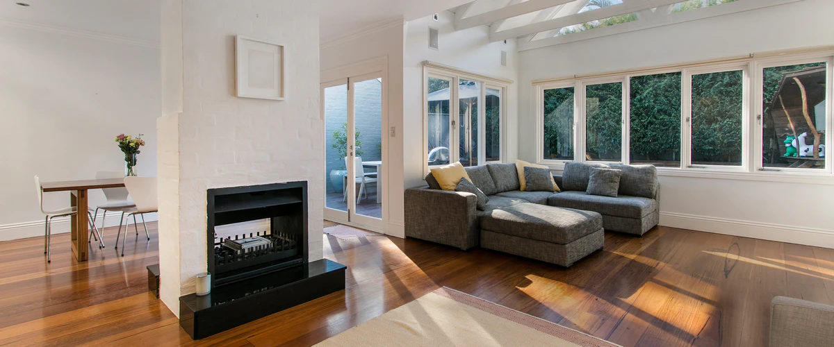 Large sunlit family room with a white brick double-sided fireplace and grey sectional sofa, demonstrating a seamless home addition.