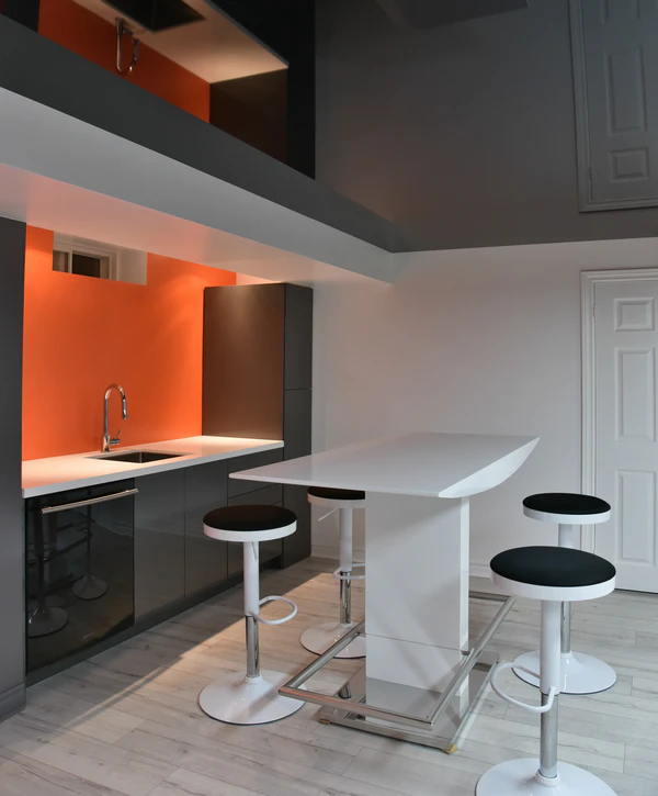 Modern basement kitchenette with an orange accent wall, dark gray cabinetry, and a white breakfast bar with stools.