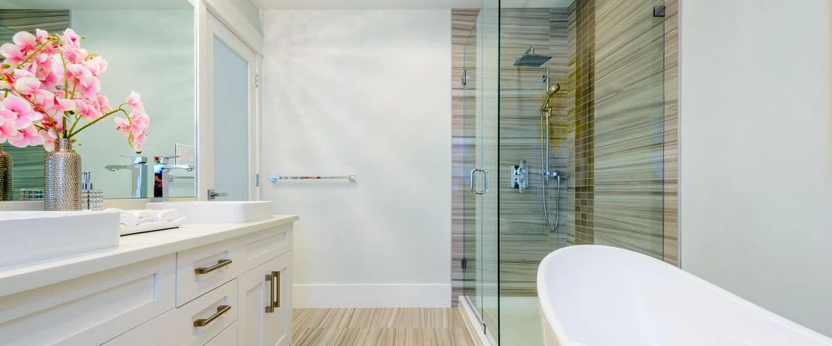 Bright modern bathroom with a white double vanity, pink flowers, a large soaking tub, and a glass-enclosed walk-in shower with neutral tile.