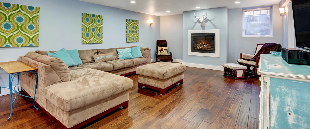 An open-concept basement family room with polished hardwood floors, light blue walls, a modern inset fireplace, and comfortable tan seating.