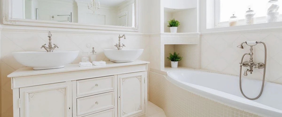 A bright white bathroom featuring a vintage-style double vanity with two vessel sinks, ornate white cabinetry, and a built-in bathtub with decorative wall niches holding greenery.
