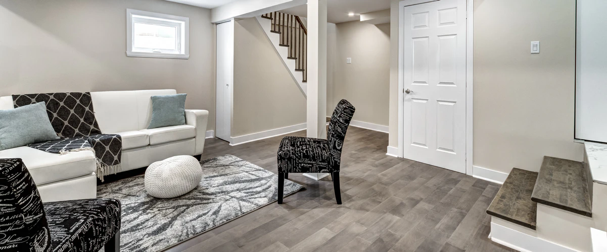 A bright, finished basement sitting area with a white sofa, script-patterned accent chair, and gray wood-look flooring near a staircase.
