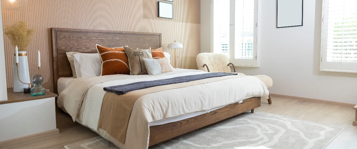 Cozy modern bedroom featuring a wooden bed frame, neutral bedding with rust orange accent pillows, a textured accent wall, and white window shutters.