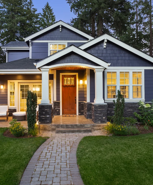 Craftsman style house exterior at dusk with dark blue siding, stone pillars, and a warm wooden front door, designed by a custom home builder in Naperville, IL.