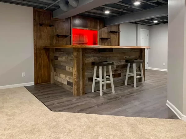 Finished basement bar with wood paneling, stone accent front, bar stools, floating shelves, and red backlit display area.